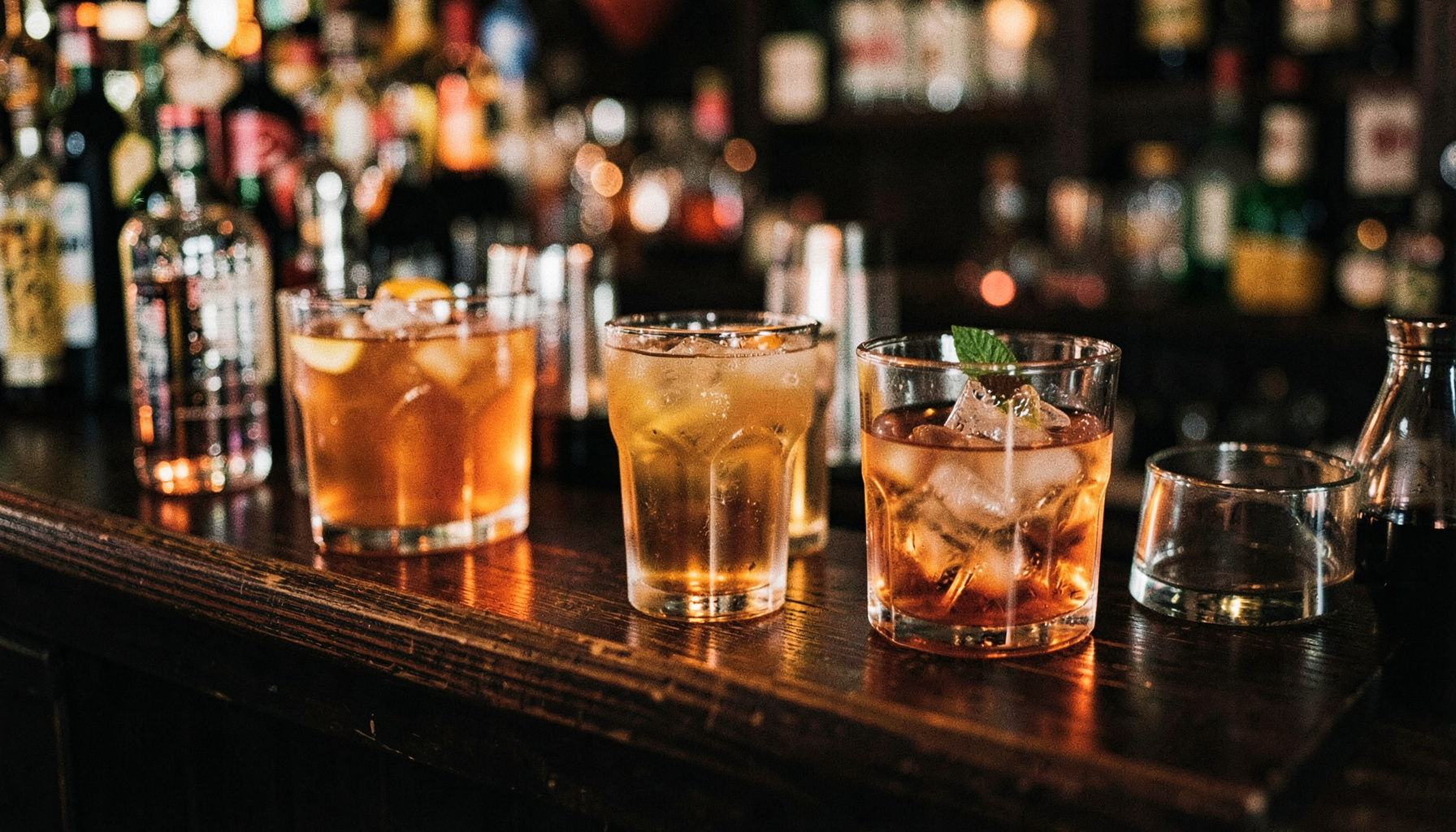 Warm amber and copper tones of cocktail ingredients on a dark wooden bar, soft moody lighting
